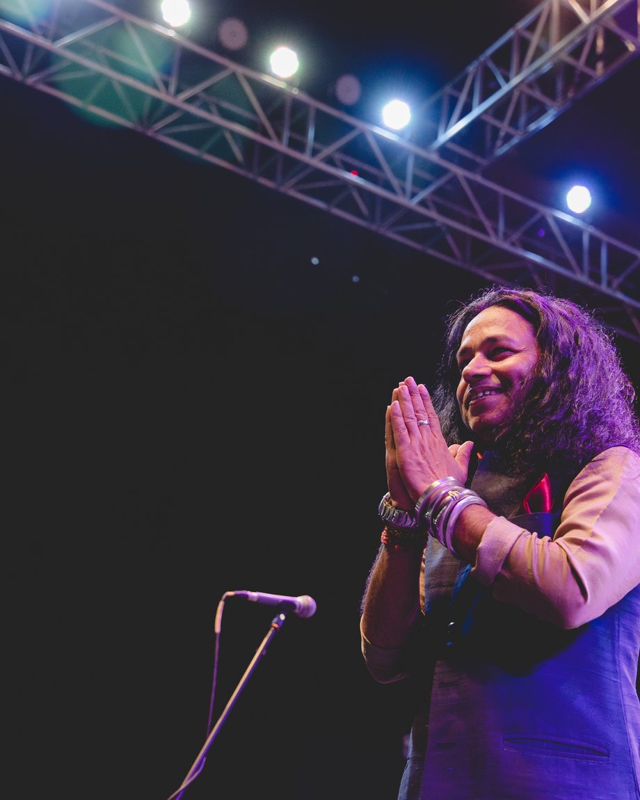 Kailash Kher greeting the audience with folded hands on stage, lighting truss overhead, at a concert produced by Outloud